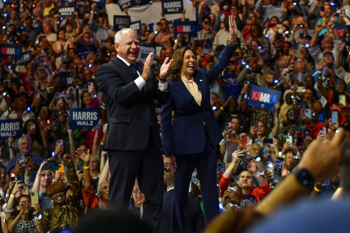 Kamala Harris and Tim Walz wave and clap in front of a large crowd.