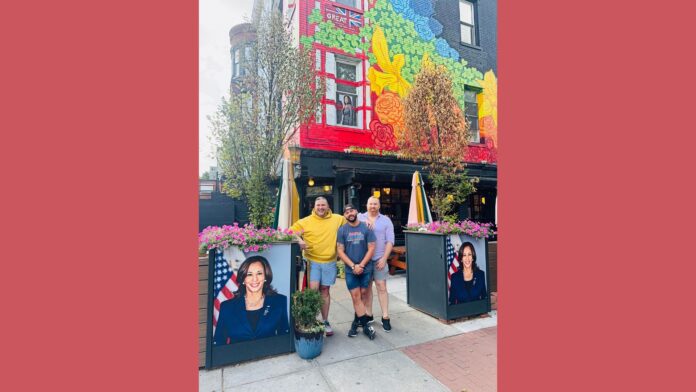 Three men pose outside a vibrant, colorful building with a large mural featuring floral designs and bright colors on its exterior. The men stand on a sidewalk next to two planters, each adorned with a large portrait of Kamala Harris. The man on the left wears a yellow hoodie, the middle man wears a blue t-shirt and black cap, and the man on the right wears a light purple shirt. Behind them, the building has a window where a person is visible looking out. The overall atmosphere is lively and celebratory.