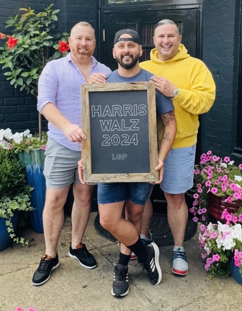 Three men stand together outside a building, smiling and holding a chalkboard-style sign that reads "Harris Walz 2024 LGP." The man on the left wears a light purple shirt, the man in the middle wears a blue t-shirt and a backward black cap, and the man on the right wears a yellow hoodie. They are surrounded by potted plants and vibrant flowers, adding a cheerful and welcoming atmosphere to the scene.