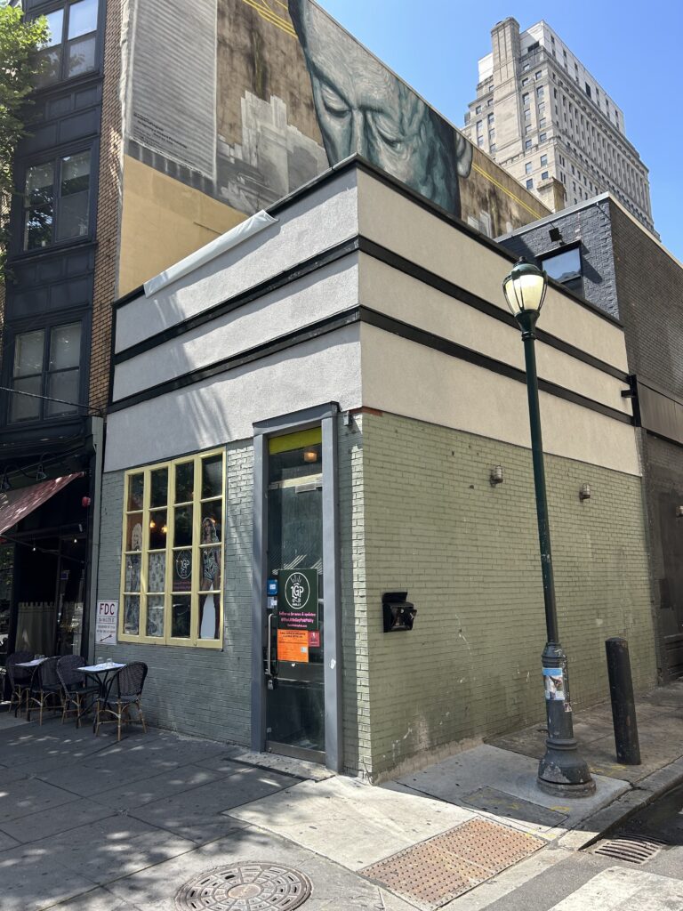 The image shows the exterior of a small building on a city street corner. The building has a light gray and greenish facade with black trim. A large mural is visible on the upper part of the building, depicting the face of a person with closed eyes in an artistic style. The door to the building has a sign that reads "Little Gay Pub," and a poster with information is displayed on the door. To the left of the door, there is a window with mannequins or posters of people visible inside. On the sidewalk outside, there is a small table with chairs, suggesting a casual dining or seating area. A streetlamp and a few trees provide shade along the sidewalk. The scene is set on a sunny day with clear skies.