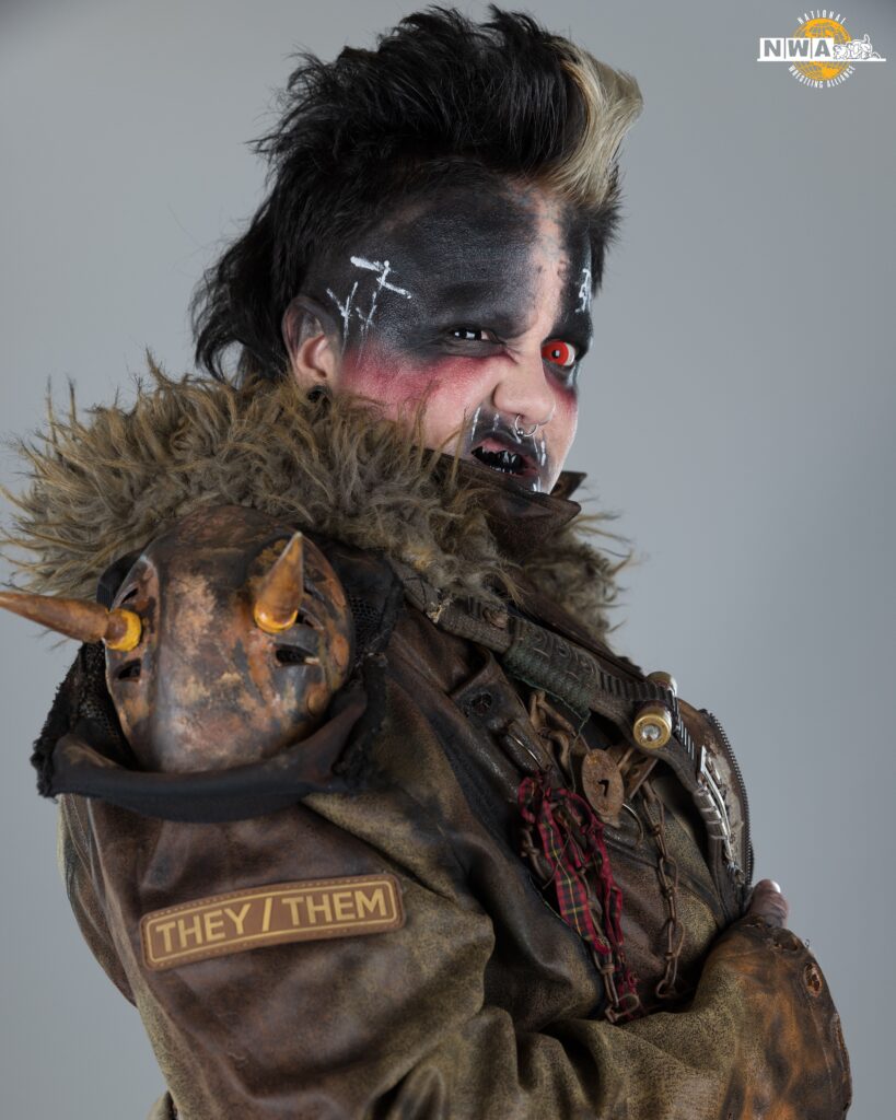 Max the Impaler, a person with an intense and unique appearance is featured in the image. They have a mohawk hairstyle with dark hair, with a blonde streak at the front. Their face is painted with dramatic black and white makeup, giving a fierce and slightly sinister look. They are wearing a rugged, post-apocalyptic style jacket adorned with various elements, including a furry collar and a small skull-like ornament with horns on the shoulder. A prominent patch on the jacket reads "THEY/THEM," indicating their preferred pronouns. The person also has piercings in their nose and wears dark, gothic-style makeup around the eyes and mouth, with one eye appearing red. The background is neutral, keeping the focus on the subject's striking appearance.