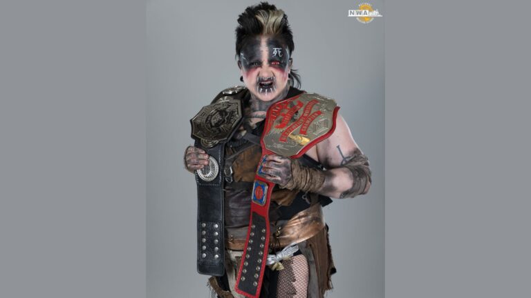 Max the Impaler is dressed in an intense, post-apocalyptic style, wearing dark makeup with black and white war paint across their face. Their hair is styled with a black and blonde mohawk. They have a fierce expression, with fangs visible in their mouth and red, glowing eyes. The person is holding two championship wrestling belts, one draped over each shoulder. Their outfit includes a mix of leather, fabric, and wraps, with tattoos visible on their arms and chest. The overall look is rugged and intimidating, with a strong warrior-like presence.