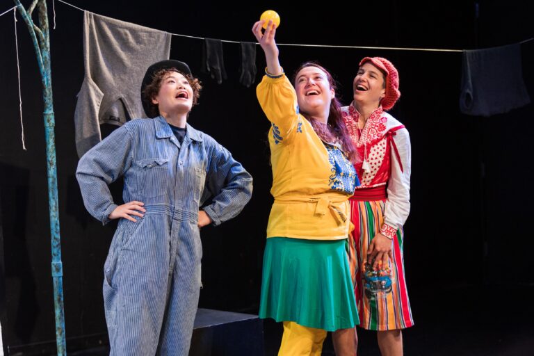 A scene from the play "War and Play," where three actors are on stage, each dressed in colorful, distinct costumes. On the left, an actor in a blue jumpsuit and black hat stands with one hand on their hip, looking upwards and appearing to sing or speak. In the center, another actor wearing a yellow top with blue embroidery, a green skirt, and yellow tights holds up a bright yellow ball, smiling as they look at it. On the right, the third actor is dressed in a red and white blouse with a red headscarf and a multicolored striped skirt, smiling at the other two. In the background, a clothesline with a gray sweater and black gloves hangs above them, suggesting a domestic or whimsical setting. The overall mood of the scene appears joyful and playful.