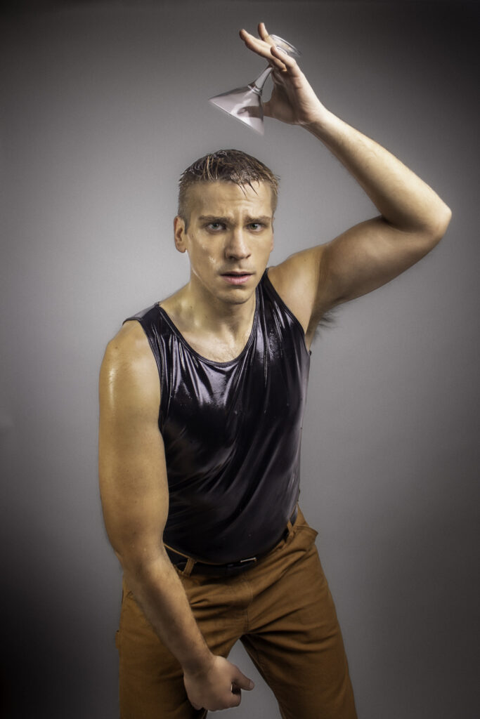 This image features Michael John Ciszewski, a performer, standing against a neutral gray background. He is wearing a sleeveless, shiny black top and light brown pants. His expression is intense and focused, with beads of sweat visible on his face and arms. He holds a martini glass upside down above his head with one hand, while his other hand is clenched in a fist, creating a dramatic and dynamic pose. The lighting highlights the contours of his muscles, and the overall mood of the image is tense and energetic.