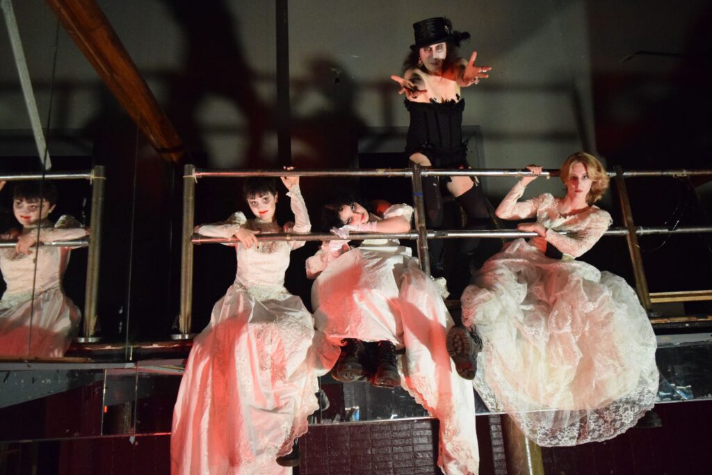 A theatrical scene from the play "My Big Fang Vampire Wedding" features four performers on a metal scaffold. Three of them, dressed in elaborate white wedding dresses with lace details, have pale faces and dramatic makeup, evoking a ghostly, vampiric appearance. They lean or sit on the scaffold, appearing languid or mysterious. The fourth performer, positioned centrally above them, wears a black corset, thigh-high stockings, and a small top hat, with exaggerated white face makeup and dark eyes, extending their arms outward in a dramatic gesture. The lighting casts ominous shadows, adding to the eerie, gothic atmosphere of the scene.