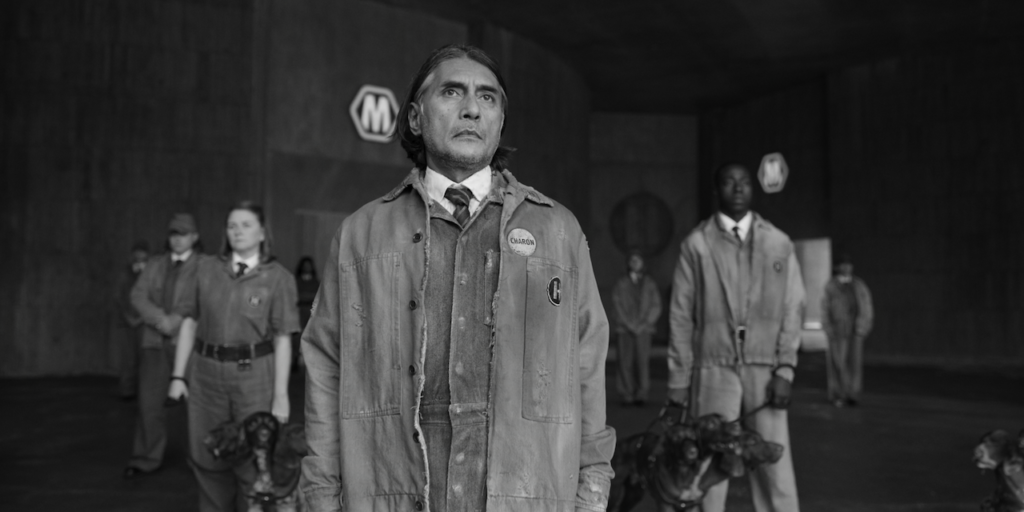 A black-and-white scene from the Netflix series Kaos featuring a group of uniformed individuals standing in a concrete, industrial-like setting. The central figure, a man with medium-length dark hair, wears a worn-out work jacket with a name tag that reads "Charon" and looks forward with a serious expression. Behind him, other people, both men and women, are wearing similar uniforms, some holding leashed dogs. The background has a large "M" logo visible on the wall, contributing to the somber and dystopian atmosphere.