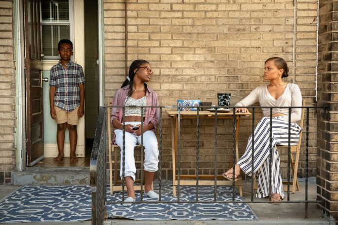 A tense scene on a small porch features three characters from Lee Daniels' "The Deliverance." A young boy stands in the doorway, wearing a plaid shirt and beige shorts, looking serious. A teenage girl sits in a chair facing sideways, wearing glasses, a pink shirt, and white pants, with a distant expression. Across from her, a woman in a light-colored top and striped pants sits at a small table, looking intently at the girl. The setting appears to be a modest, brick-walled home. The mood is somber and reflective.