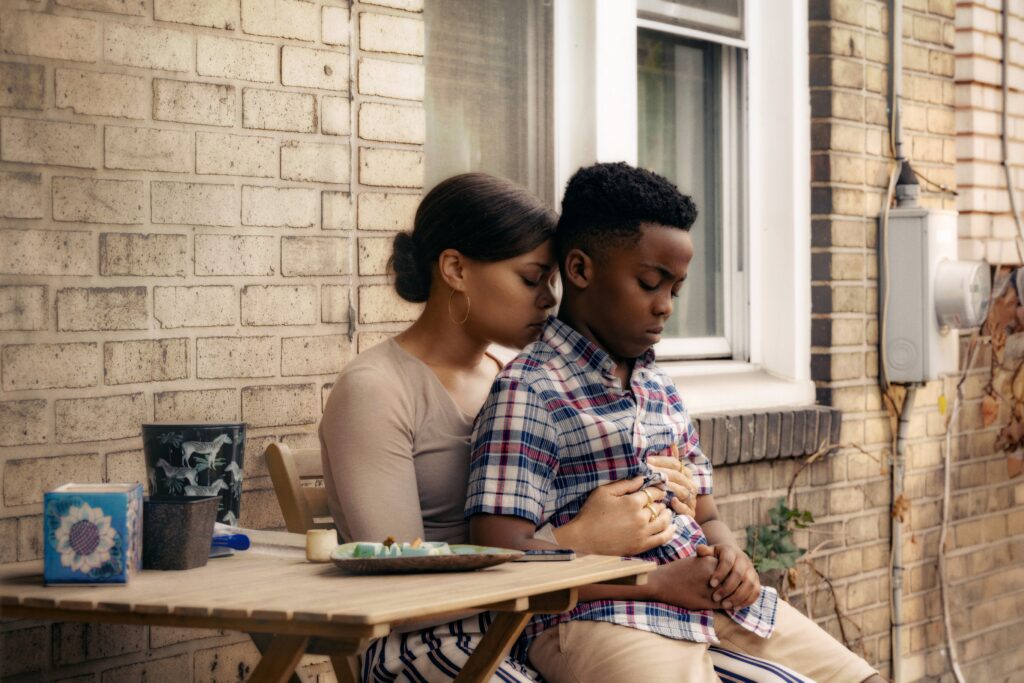 In an emotional scene from "The Deliverance," a woman tenderly embraces a young boy on a small porch. The woman, wearing a light-colored top and hoop earrings, rests her head against the boy's as she holds him tightly, conveying a sense of deep concern and affection. The boy, dressed in a plaid shirt, sits quietly with a somber expression, leaning into the woman's embrace. A small table beside them holds a few objects, including a plate with food and a cup, adding to the intimate atmosphere. The brick wall of the house serves as a backdrop to this poignant moment.