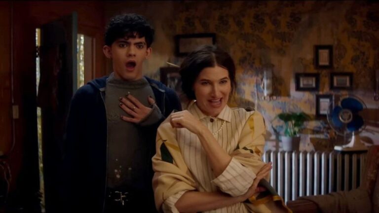 Joe Locke, looking surprised with one hand on his chest, stands next to a confident and playful Kathryn Hahn, arms crossed, in a scene from Agatha All Along. They're in a cozy, vintage-style room with floral wallpaper, plants, and framed pictures, capturing a lighthearted moment between their characters.