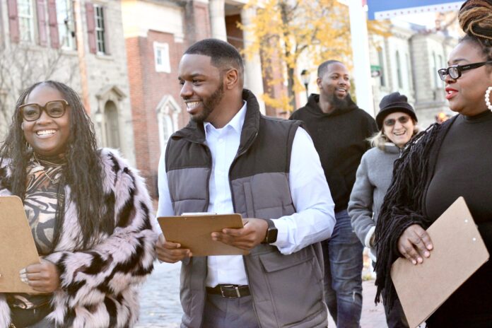 A group of people, including Andre Carroll (center), walk together outdoors in a friendly and lively atmosphere. Andre, wearing a light blue shirt and a gray vest, smiles while holding a clipboard. He is accompanied by two women on either side, both holding clipboards, with one woman wearing a fur-style coat and large glasses. In the background, additional people join the group, one of them laughing, and a scenic backdrop of brick buildings and fall foliage enhances the image.