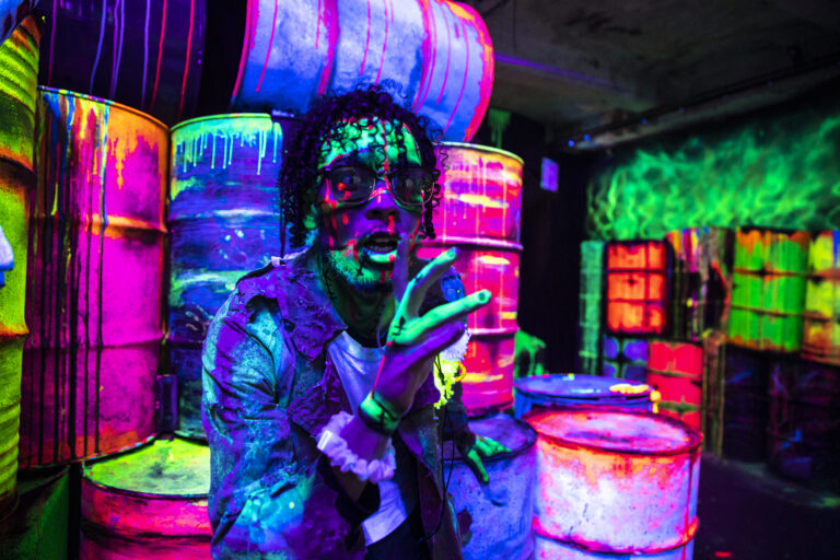 Anthony Salas, dressed as a zombie with green and purple face paint, stares intensely at the camera in a dark, neon-lit setting. He stands in front of colorful, glowing barrels, creating a vibrant, eerie atmosphere.