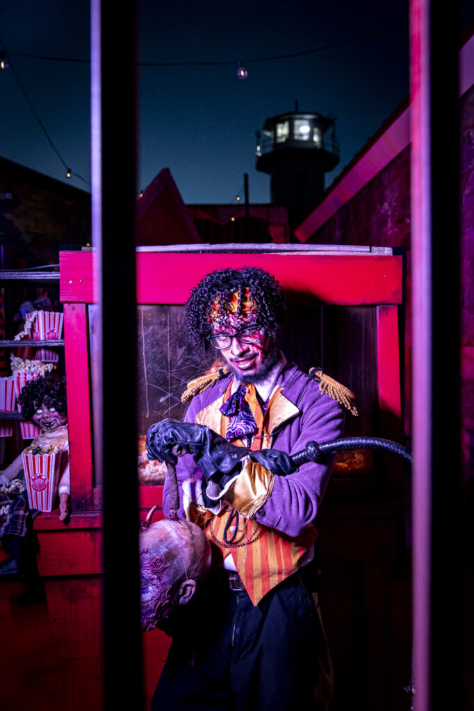 Anthony Salas, dressed in a ringmaster costume with gory face paint, stands holding a prop severed head and whip in a spooky, carnival-themed setting. Behind him, a creepy doll sits among popcorn containers, adding to the eerie atmosphere.