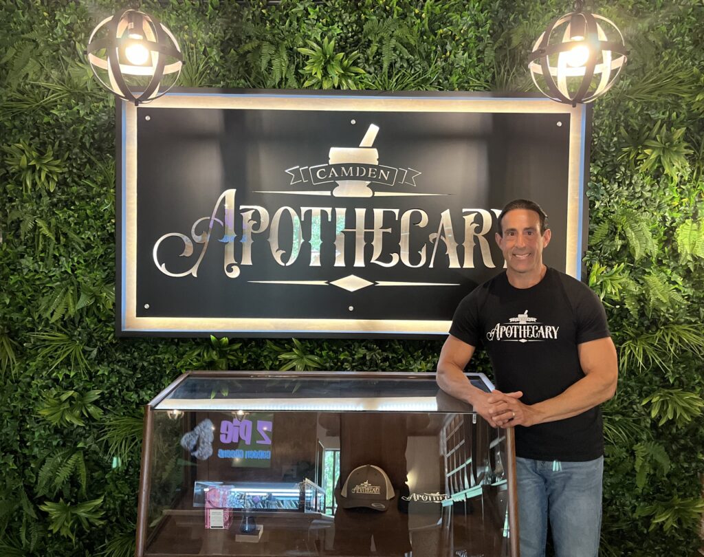 Tony Minniti stands in front of a display case and a large sign that reads 'Camden Apothecary.' The backdrop features a lush green wall with plants, creating a natural atmosphere. The man is smiling and wearing a black T-shirt that also says 'Camden Apothecary.' The display case in front of him contains merchandise, including a hat with the same logo.