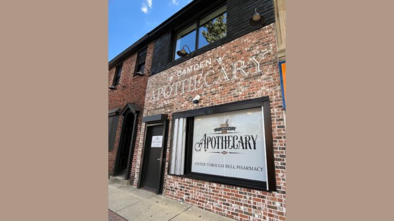 A brick building with a vintage-style sign that reads 'Camden Apothecary' in large white letters. The facade features black trim around the windows and doors. The entrance is labeled 'Enter Through Bell Pharmacy' on a large window display, with a smaller door to the left marked 'Exit Only.' The building's exterior combines old-world charm with a modern touch.