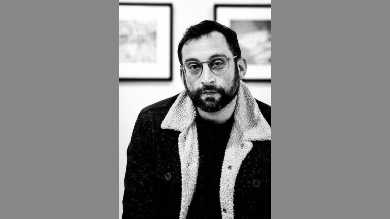 A black-and-white headshot of Cory David Botnicker. He has a short beard, wears round glasses, and looks directly into the camera with a calm expression. He is dressed in a jacket with a soft shearling collar. The background features framed artwork, slightly out of focus.