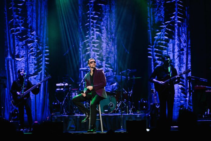 Craig Winberry performs in The Life and Music of George Michael. He sits confidently on a stool in the center of the stage, holding a microphone, wearing sunglasses and a dark blazer. The stage is illuminated by blue and green lights, with intricate background drapery. A bassist and guitarist accompany him in the dimly lit background, surrounded by various musical equipment and instruments. The atmosphere evokes a dynamic and moody concert setting, highlighting the tribute to George Michael's iconic music.