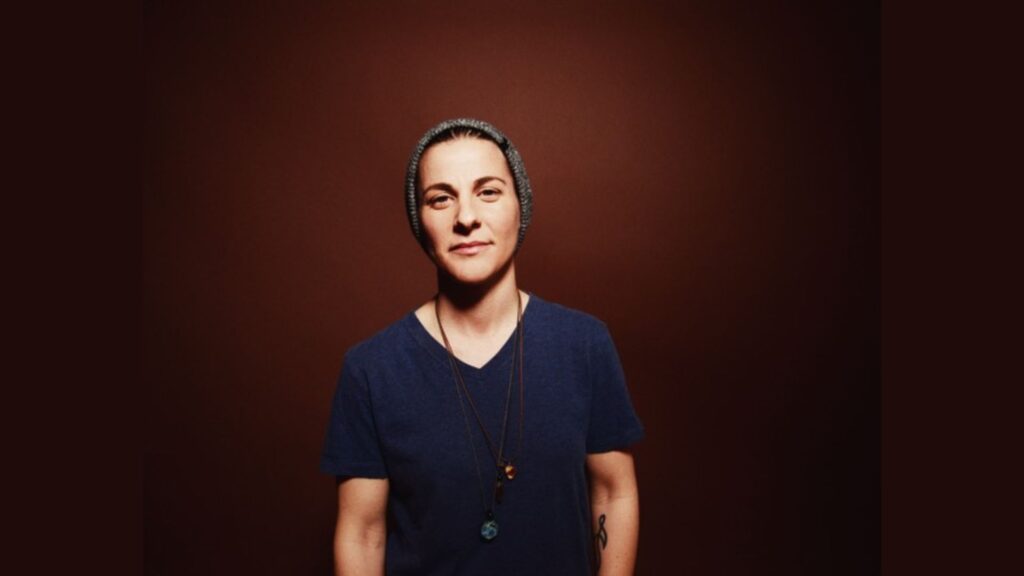 This image features Andrea Nardello standing in front of a plain brown background. Andrea is wearing a gray beanie, a dark blue T-shirt, and two necklaces. The expression is calm and confident, with hands relaxed by the sides. A small tattoo is visible on the left forearm. The lighting is soft, casting subtle shadows, and emphasizing the simplicity and focus on Andrea's presence.