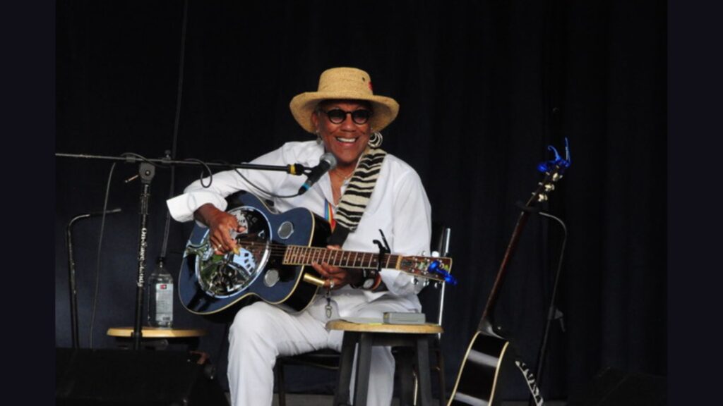 This image shows Gaye Adegbalola seated on stage, smiling while playing a resonator guitar. She is wearing a straw hat, round dark sunglasses, and a white outfit, accessorized with large hoop earrings and a zebra-patterned scarf draped over her shoulder. The background is dark, with microphones and a second guitar visible, giving the scene a warm and intimate performance atmosphere.