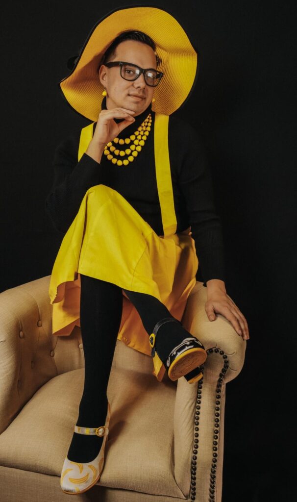 Jacob Kelly is seated on a beige chair, dressed in a vibrant yellow ensemble. They wear a wide-brimmed hat, a bold yellow necklace, and matching earrings. Their outfit includes a yellow suspender skirt paired with black tights and shoes featuring yellow accents. They strike an elegant pose with one hand resting under their chin.