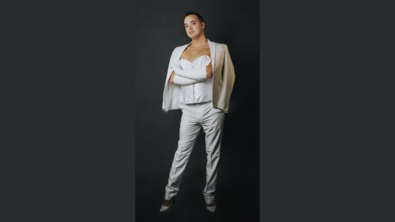 Jacob Kelly stands confidently in an all-white outfit, combining a sleek white suit with a structured corset-style top and long gloves. The look is completed with high heels, showcasing a stylish blend of traditionally feminine and masculine elements.