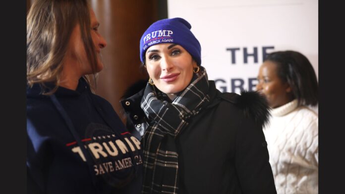 Laura Loomer wearing a blue beanie with the slogan "TRUMP Make America Great Again!" written on it. She is dressed in a black coat with a black-and-white plaid scarf. Her expression is neutral, and she is standing close to another person wearing a hoodie with a Trump slogan. Another individual in the background appears slightly out of focus.