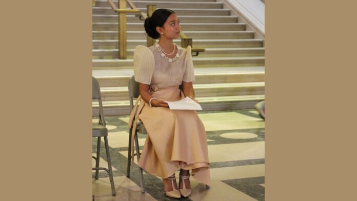 Malaya Ulan sits on a chair in front of a staircase, dressed in a formal beige skirt and top with puffed sleeves. She holds a sheet of paper in her hands, and her hair is neatly styled in a bun. The setting appears to be indoors, with marble floors and grand staircases in the background. She looks attentive and poised, her gaze directed slightly to her right. The atmosphere suggests a formal or cultural event.