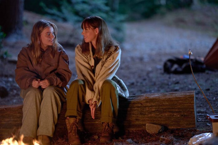 From left, Maisy Stella and Aubrey Plaza sit on a log in the woods at night in My Old Ass, sharing a warm conversation by the fire. Both are dressed casually, with Maisy in a large jacket and Aubrey wrapped in a cozy sweater, creating a calm and intimate moment in the outdoors.