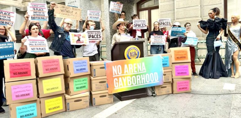 Yoojin Park stands among more than a dozen files boxes and behind a podium. He wears a cowboy hat. More than two dozen protesters stand behind him with signs supporting the No Arena movement.