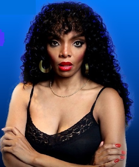 A portrait of Raniere Martin, a Donna Summer tribute singer, standing confidently against a blue background. She has voluminous curly hair and wears bold red lipstick. Her black lace top reveals her strong presence, and she accessorizes with large hoop earrings and a delicate gold necklace. Her arms are crossed, exuding a sense of strength and poise, with a serious expression that captures her intensity and focus.