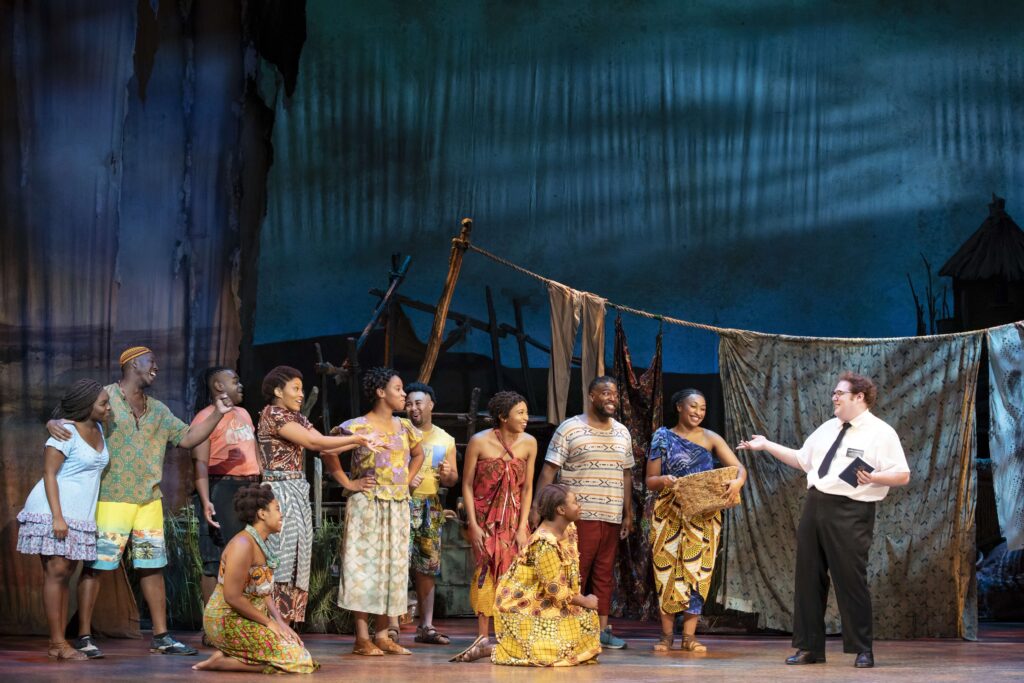 This is a scene from the musical The Book of Mormon, set in an African village. The cast members on stage wear brightly patterned traditional African clothing. Most of them are smiling and engaging in conversation. One character, dressed in a white shirt, black tie, and black pants, typical of a Mormon missionary, is speaking to the villagers while holding a book, likely a reference to the Book of Mormon. The background features a rustic village with simple structures, drying clothes on a line, and earthy, natural tones