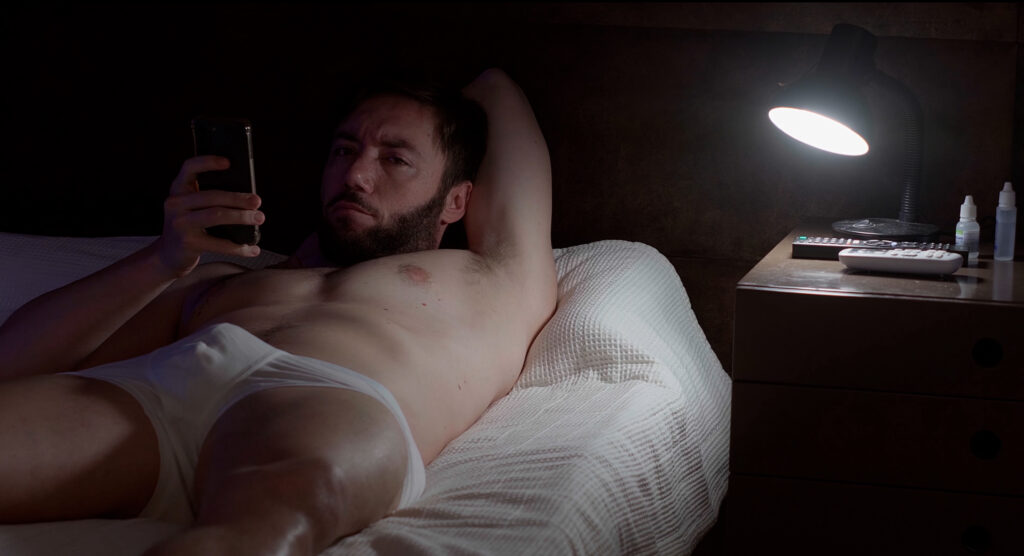 This image shows Sergio Harger in a scene from "The Senator," lying on a bed, partially covered by a white blanket. He is shirtless and wearing only white briefs, resting with one arm behind his head while holding a phone in his other hand. His expression appears serious and contemplative, as he gazes at the screen. The dimly lit room is illuminated by a bright bedside lamp, casting light over a small nightstand, which holds several objects, including a remote control, a bottle of eye drops, and a small container.