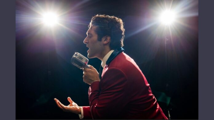 Will Stephan Connell, dressed as Frankie Valli in a vibrant red suit with a white shirt and dark collar, passionately sings into a vintage silver microphone under bright stage lights. His hand is gesturing expressively as he performs, embodying the essence of the iconic 'Jersey Boys' character.