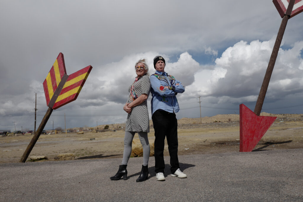 Two individuals are standing back to back in a barren landscape with large, colorful arrows pointing toward the ground on either side of them. Harper Steele, on the left, is wearing a patterned dress, gray leggings, black heeled boots, and tinted sunglasses. Will Ferrell, on the right, sports a light blue jacket with colorful patches, black pants, a beanie, and sneakers. Both strike confident, playful poses against a cloudy sky and a desolate desert setting.