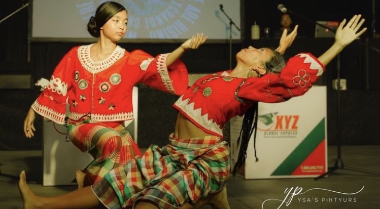 Malaya Ulan and her mother, Annielille (Anito) Gavinocan, perform a graceful dance on stage, both wearing matching red traditional attire with intricate embroidery. Malaya stands with her arm outstretched, her expression focused and calm, while Annielille leans back dramatically, balancing on one knee with her arms extended, her braided hair flowing behind her. The background shows a banner and microphones, indicating a formal or cultural event. Their synchronized movements reflect deep cultural significance and artistic expression.