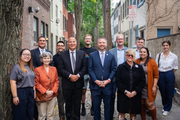 Twelve people are pictured on a cobblestone street in Phialadelphia. They stand as a group and look at the camera smiling.