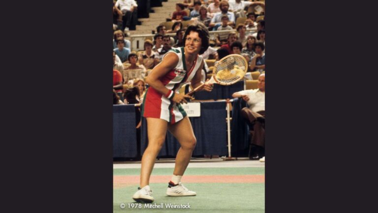 Billie Jean King is shown on the court, wearing a colorful striped dress and holding a wooden racket, ready to return a shot. Her short dark hair, glasses, and confident stance are iconic of her 1970s era, with the background featuring a lively crowd of spectators.