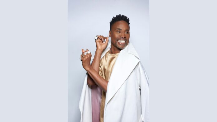 Billy Porter smiling brightly, wearing a beige satin top with a white coat draped over his shoulders. He has short, styled hair and is showcasing several large silver rings on both hands, which are raised near his face in a playful pose.