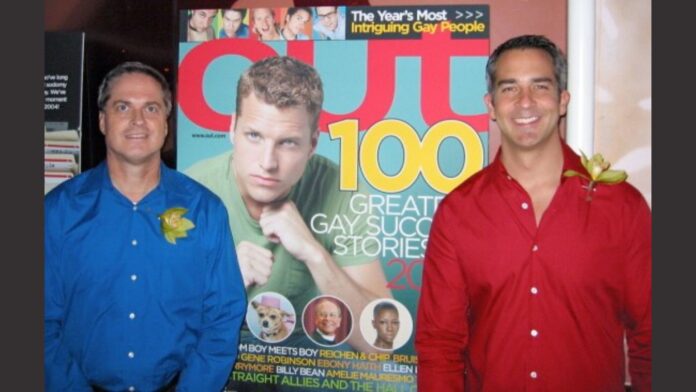 Bob Witeck (left) and Wes Combs (right) standing in front of an Out magazine cover featuring the "100 Greatest Gay Success Stories" from 2003. Witeck is wearing a blue shirt with a yellow ribbon, while Combs is wearing a red shirt with a matching ribbon.