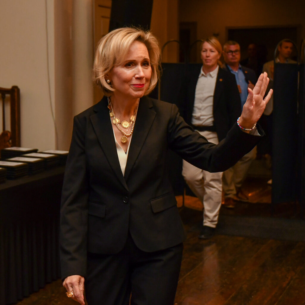 Gwen Walz, dressed in a black suit with a white blouse, is smiling and raising her hand in a greeting or wave. She is wearing layered gold necklaces and earrings. In the background, several people, including security personnel, are following her in what appears to be a formal event setting.