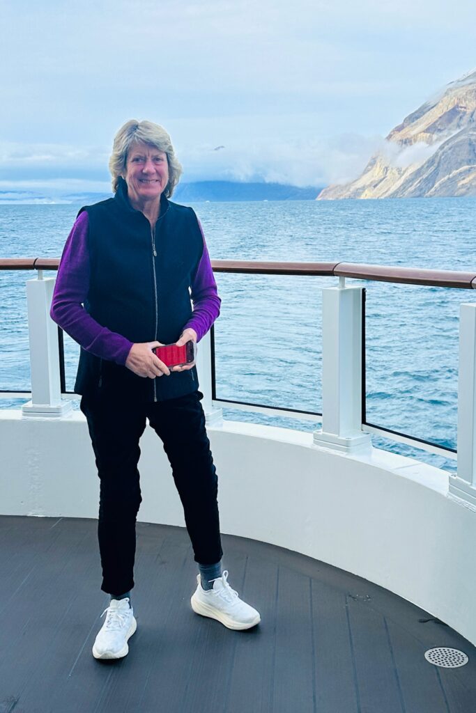 Marianne Lieberman stands on the deck of a boat, smiling and holding a red phone, with the calm sea and a mountainous landscape in the background. She is dressed in a black vest, purple sweater, black pants, and white sneakers, enjoying the serene ocean view.