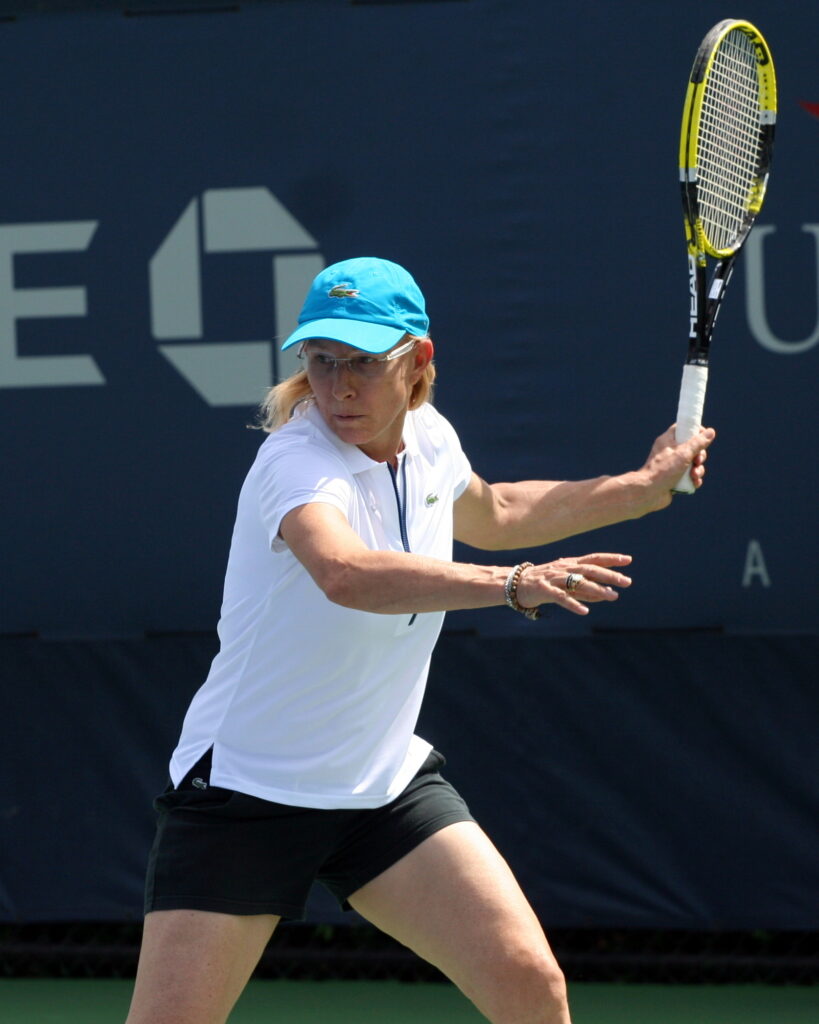 Martina Navratilova is captured in action, wearing a white top, black shorts, and a blue cap. Her intense focus is evident as she prepares to hit the ball with a modern racket, set against the backdrop of a professional tennis court.