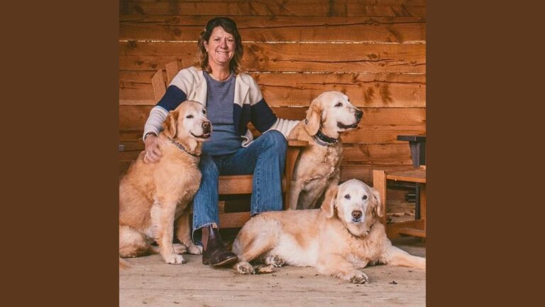 Marianne Lieberman sits in a wooden chair, smiling warmly, surrounded by three golden retrievers. The cozy setting features wooden paneling in the background, and the dogs are relaxed, with one resting on the floor while the others sit beside her.