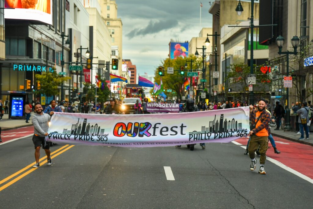 Participants in the OURfest Philly Pride 365 parade carry a large banner down a city street, celebrating National Coming Out Day. The colorful scene features people holding pride flags, while onlookers gather along the sidewalks in a bustling downtown area, surrounded by tall buildings and bright signs.