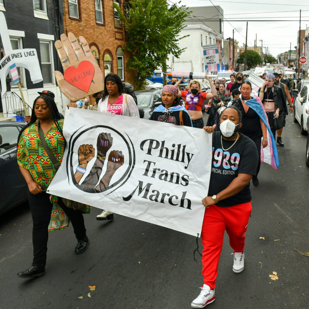 A group of people are marching down a street holding a banner that reads "Philly Trans March" with three raised fists of different skin tones depicted on it. Some marchers are wearing face masks, and one individual at the front is holding a large cutout of a hand with the word "Healing" written inside a heart on the palm. Many participants are dressed in vibrant clothing and carrying transgender pride flags. The background shows a neighborhood with row houses and parked cars along the street. The overall atmosphere suggests a peaceful protest or community event.