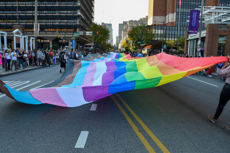 A large crowd gathers in the streets of Philadelphia, holding a vibrant, extended Progress Pride flag. The flag stretches across the road, featuring colors representing the LGBTQ+ community, including the rainbow, trans pride, and black and brown stripes. People along the sidewalk watch the event, some wearing Pride-themed apparel. The scene takes place in the heart of the city, with tall buildings and Independence Visitor Center visible in the background, capturing the spirit of celebration and inclusivity.