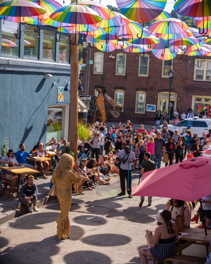 A vibrant outdoor event is taking place under a colorful canopy of rainbow umbrellas. A performer in a shimmering gold outfit stands on a small street stage, addressing an enthusiastic crowd of onlookers seated and standing along the sidewalk. The audience includes people holding small pride flags, showing their support. Nearby, patrons sit at tables beneath umbrellas enjoying the event. The scene is set in a lively neighborhood, with brick buildings in the background and festive decorations adding to the atmosphere of celebration and inclusivity at this "Out & About in MNYK" event.