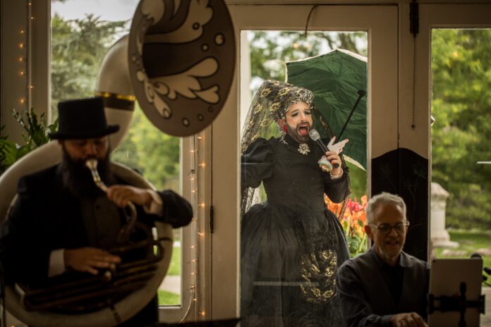 The Bearded Ladies Cabaret performs at the Laurel Hill Cemetery Picnic in 2023. In the foreground, Josh Machiz plays the sousaphone, while Jarbeaux, wearing an elaborate black dress with a lace veil and holding a green umbrella, sings into a microphone outside the window. . Heath Allen plays keyboard, smiling, in the lower right corner of the image. The scene takes place against a backdrop of greenery and natural light entering through the windows."