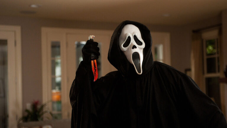 The image shows a figure wearing a black hooded robe and the iconic white Ghostface mask, commonly associated with the "Scream" movie franchise. The figure holds a knife dripping with red liquid, which resembles blood, creating a menacing and suspenseful atmosphere. The background suggests an indoor setting, with large windows and soft lighting, giving the scene a mix of everyday normalcy and horror.