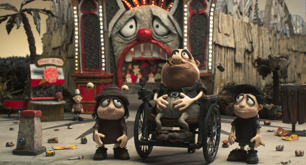 A scene from the animated film "Memoir of a Snail," which is screening at the Philadelphia Film Festival, features three claymation characters standing in front of a run-down amusement park. The character in the center, a man in a wheelchair, has a slouched posture, a cigarette in his mouth, and a melancholic expression. To his left is a smaller character with large eyes, wearing a hat with an antenna, and to his right is a young boy with spiky hair, looking up with a sad, hopeful expression. The background shows a tattered carnival booth labeled "Fairy Floss" and a large, clown-faced entrance with glowing lights. Scattered trash litters the ground.