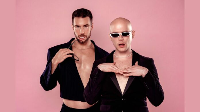 In this promotional image for Mean Gays, Jake Jonez (left) and Aaron Goldenberg (right) are posing against a pink background. Jake has short dark hair, a trimmed beard, and is wearing a partially open, cropped black jacket, holding sunglasses to his lips. Aaron is bald, wearing white-framed sunglasses and a black jacket, with a string of pearls around his neck. Both are dressed in an edgy, fashion-forward style, exuding confidence and attitude. Their expressions and poses suggest a bold, playful vibe, perfectly aligned with the campy and irreverent tone of the show.